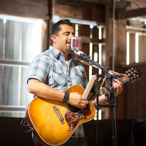 A man in a plaid shirt sings into a vintage microphone while playing an acoustic guitar.