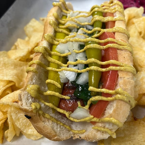 A hot dog in a bun with mustard, relish, onions, tomatoes, and peppers, served with chips.