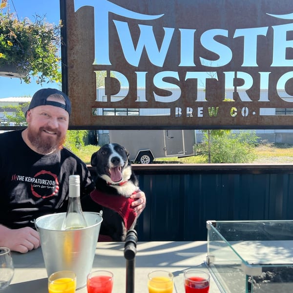 Man and dog smiling at an outdoor table with drinks and a Twisted District Brew Co. sign.
