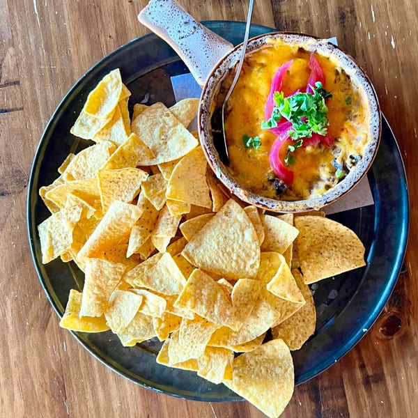 A bowl of melted cheese dip with pickled onions and cilantro, served with tortilla chips on a dark plate.